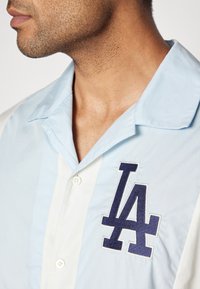 Hellblaues und weißes Kurzarmhemd mit einem gestickten "LA"-Logo in Dunkelblau. Mit Knopfverschluss und einer glatten, gewebten Textur.
