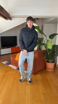 Νέος άνδρας που φοράει μαύρο μπουφάν και ανοιχτό μπλε τζιν στέκεται με τα χέρια ενωμένα σε ένα σύγχρονο σαλόνι με φυτά και τηλεόραση.