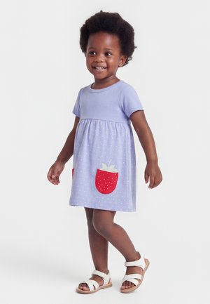 Tout-petit souriant portant une robe violet clair avec des poches rouges en forme de fraises et des sandales blanches, debout devant un fond uni.