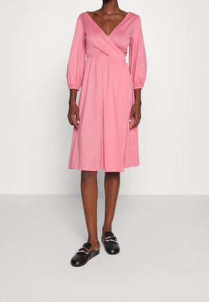 Mujer que lleva un vestido cruzado rosa hasta la rodilla con mangas abullonadas tres cuartos y zapatos negros tipo mocasín sin cordones con hebillas plateadas.