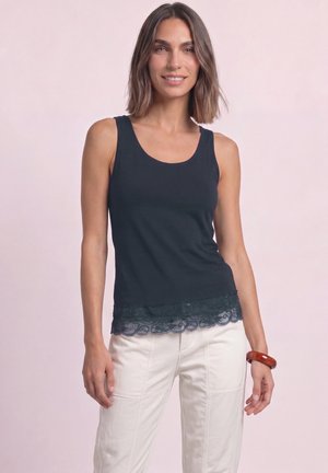 Femme portant un haut sans manches noir avec un ourlet en dentelle et un pantalon blanc, debout devant un fond clair uni, souriant légèrement.