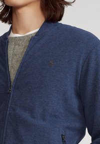 Giacca zip-up blu realizzata in tessuto morbido, con uno strato interno grigio testurizzato e un piccolo logo ricamato sul petto.