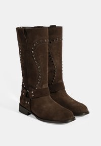Bottes en daim marron avec un design épuré, des accents cloutés sur les côtés et une sangle décorative autour de la cheville. Semelles basses et plates.