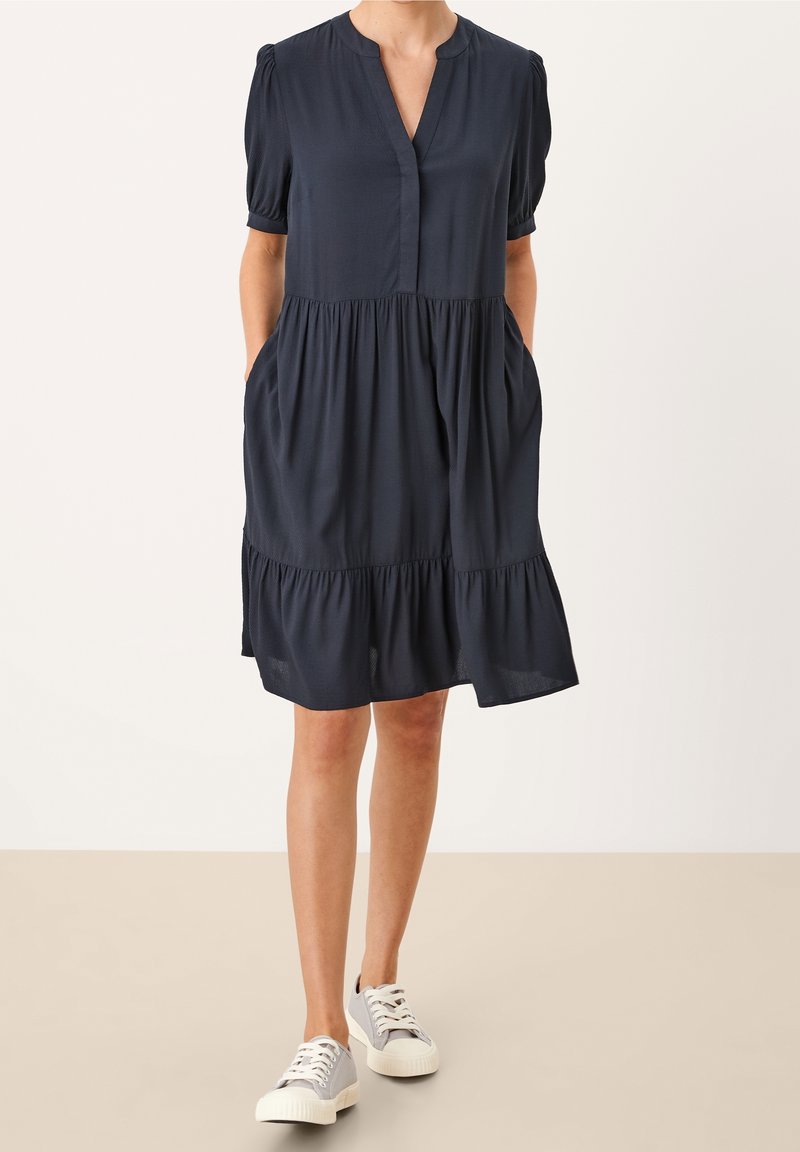 Femme portant une robe bleu marine fluide à manches courtes avec une jupe à volants et des baskets blanches, debout les mains dans les poches sur un fond neutre.