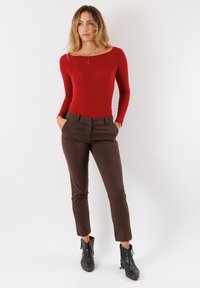 Body a maniche lunghe rosso abbinato a pantaloni marroni sagomati e stivali neri alla caviglia. Tessuto liscio, vestibilità aderente e tasche funzionali visibili.