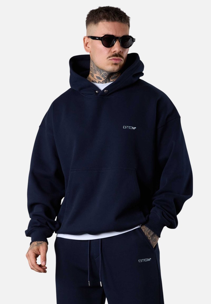 Un sweat à capuche bleu marine en tissu doux présente une poche kangourou et un logo brodé sur la poitrine. Le modèle porte des lunettes de soleil noires.