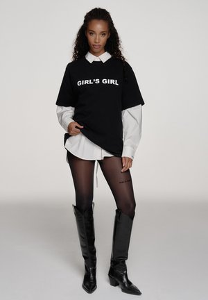 Vrouw staat en draagt een zwart shirt met de tekst "GIRL'S GIRL" over een wit shirt, doorschijnende zwarte panty met tekst en zwarte leren laarzen tot aan de knie.