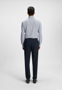 Ljusblå mönsterfylld skjorta med spetsig krage, i kombination med mörka marinblå byxor och svarta skor, sedd bakifrån.