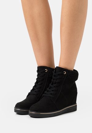 Bottines compensées noires en daim avec un col texturé. Elles présentent des détails en métal doré, un lacet et une semelle en caoutchouc avec une semelle extérieure distincte.