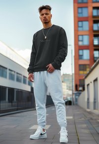 Zwarte sweatshirt met witte logo, gecombineerd met lichtblauwe joggers. Persoon draagt witte sneakers, heeft een kort kapsel en een ketting.