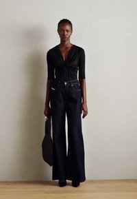Svart langermet topp med knyttet front, kombinert med vide, mørke denimbukser. Modellen holder en svart veske, stående mot en nøytral bakgrunn.