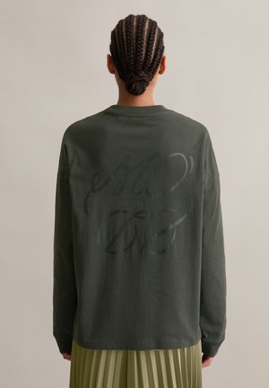Person mit geflochtenem Haar in einem tiefen Dutt, trägt ein dunkelgrünes Langarmshirt mit schwarzem geschwungenem Text auf dem Rücken und einen hellgrünen plissierten Rock.