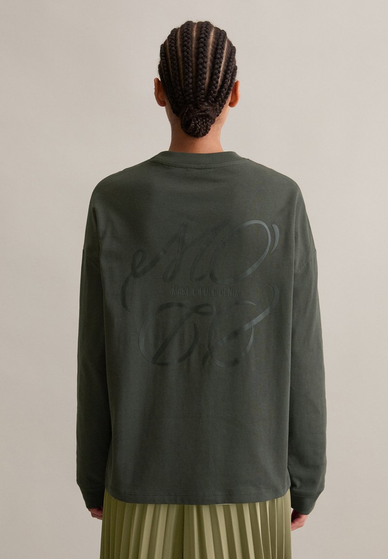 Persona con capelli intrecciati in un basso chignon, indossa una maglietta a maniche lunghe verde scuro con testo corsivo nero sulla schiena e una gonna plissettata verde chiaro.