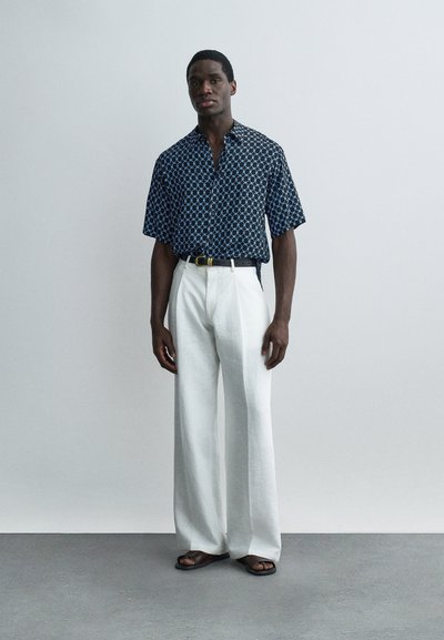 Άνδρας που στέκεται μπροστά σε έναν απλό τοίχο, φορώντας μπλε σκούρο πουκάμισο με σχέδια και κοντά μανίκια, άσπρο παντελόνι με φαρδιές παντελόνες, μαύρη ζώνη και μαύρα σανδάλια.