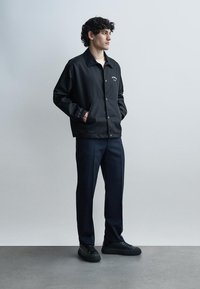 Jeune homme aux cheveux bouclés portant une veste noire boutonnée, un pantalon foncé et des baskets noires, debout les mains dans les poches sur un fond uni.