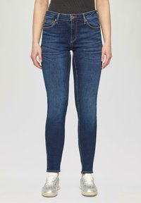 Jean slim en denim bleu foncé, avec une taille haute, des coutures orange et des poches avant. Accompagné de baskets argentées.