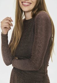 Long-sleeve top in dark brown metallic fabric, featuring a fitted design with an even texture and subtle shimmer. Arm crossed.