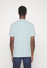 Světle modré tričko s krátkými rukávy a kulatým výstřihem, vyrobené z měkké, hladké tkaniny, s viditelnou nášivkou loga na boku.