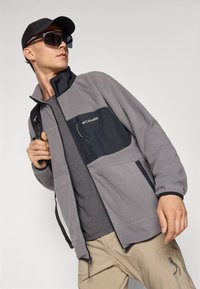 Giacca in pile grigia con una tasca sul petto in nylon nero e cerniera. Abbinata a una t-shirt grigia e pantaloni beige. Un cappello nero e occhiali da sole completano il look.
