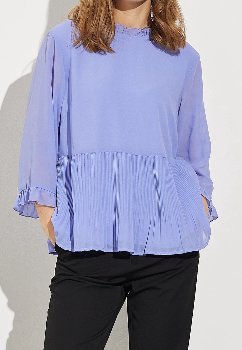 Lavendelfärgad plisserad blus med korta volangärmar och lös passform. Tyget är lätt och halvgenomskinligt, med vertikala veck vid nederkanten.