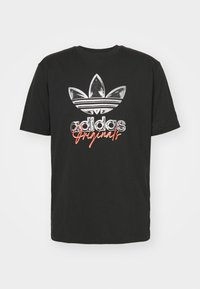 Černé bavlněné tričko s velkým stříbrným logem Adidas trefoil a červeným textem "Originals". Kulatý výstřih a krátké rukávy. Klasický střih.