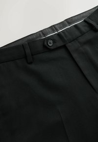 Pantalon de costume noir avec bouton à la taille, passants de ceinture et doublure intérieure visible sur un fond clair.