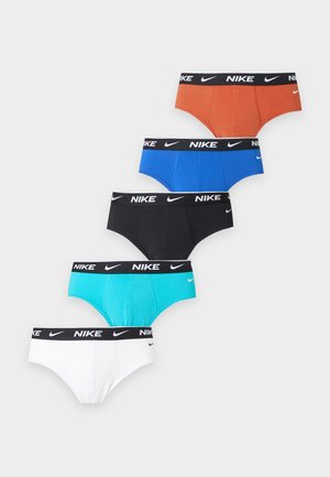 Ung sort mand med en veltrænet fysik iført orange Nike underbukser, stående barfodet mod en ensfarvet hvid baggrund og kigger til siden.