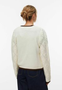Pull en maille blanche avec des manches texturées à motif losange, des poignets côtelés marron, un col arrondi et une coupe décontractée.