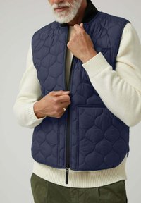 Chaleco acolchado azul marino con un patrón texturizado, cremallera frontal y dos bolsillos. Superpuesto sobre un suéter crema con puños acanalados.