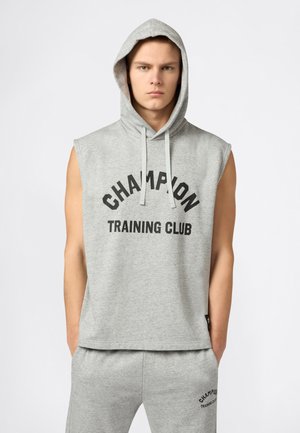 Jonge man die een grijze mouwloze hoodie en bijpassende joggingbroek draagt met de tekst "Champion Training Club" aan de voorkant.