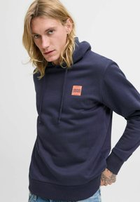 Navy blue hoodie in soft fabric, featuring a front pocket, drawstrings, and a small orange logo on the chest. Textured cuffs and hem.