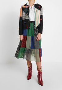 Veste en patchwork avec des motifs multicolores, blouse noire, et jupe superposée présentant diverses couleurs et imprimés. Bottes de cow-boy rouges avec des accents.