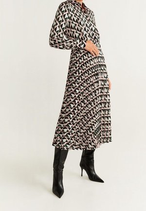 Kobieta ubrana w sukienkę midi z długim rękawem o geometrycznym wzorze i plisowaną spódnicą, w połączeniu z czarnymi, szpiczastymi botkami na wysokim obcasie.