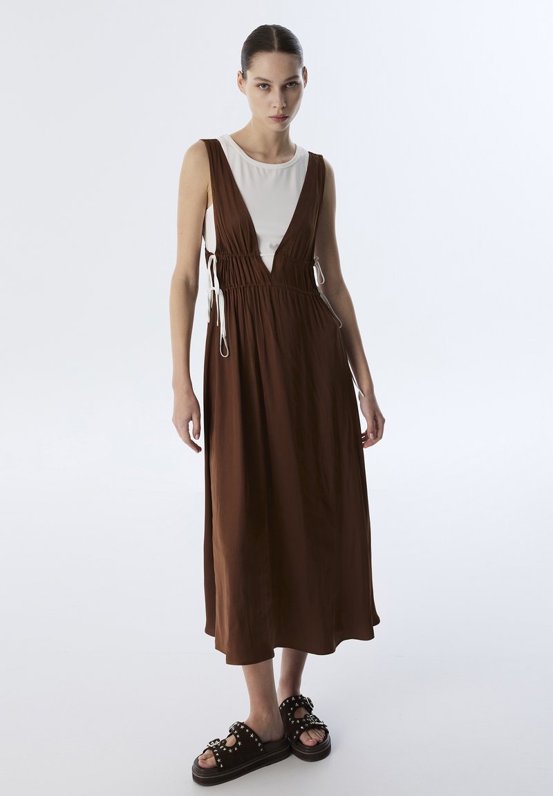 Bordeaux zonder mouwen jurk met een diepe V-hals, gathered taille, wit binnenhemd, en zwarte sandalen met banden en studaccents.