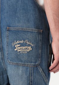 Denim tuinbroeken in vervaagd blauw, met een achterzak waarop het witte geborduurde "Tommy Jeans" logo staat. Contrasterende stiksels zichtbaar op de randen.