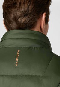 Zielona kurtka puchowa z wysokim kołnierzem, z poziomym pikowaniem i pomarańczowym logo "HACKETT" blisko kołnierza.