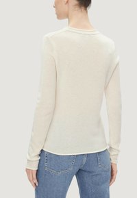 Donna vista di spalle che indossa un maglione a maglia beige a maniche lunghe e jeans in denim blu, su uno sfondo chiaro uniforme.