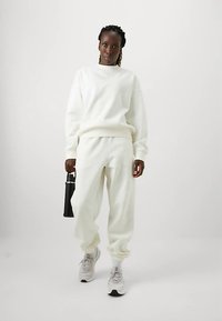 Biała polarowa bluza i dopasowane dresy z prążkowanymi mankietami. W połączeniu z jasnoszarymi trampkami. Trzyma czarną butelkę. Prosty, czysty design.