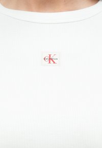 Balta ribota krekla apkakle ar nelielu taisnstūra formas Calvin Klein logotipa plāksteri ar sarkanām "CK" burtiem.