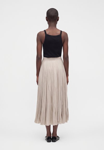 Femme debout de dos portant un débardeur noir, une jupe midi plissée beige et des chaussures plates noires sur un fond uni.