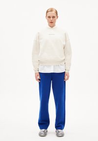 Lichtbeige oversized sweatshirt met ribbelmanchetten, gecombineerd met felblauwe trainingsbroek. Model draagt zilveren sportschoenen. Witte achtergrond.