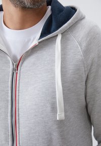 Grijze zip-up hoodie met een marineblauwe gevoerde capuchon, witte stropdassen en een subtiele rode en marineblauwe streep langs de rits. Zachte stoffen textuur.