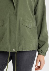 Giacca leggera verde oliva in tessuto liscio, dotata di due tasche sul petto, polsini elastici e orlo con cordoncino.
