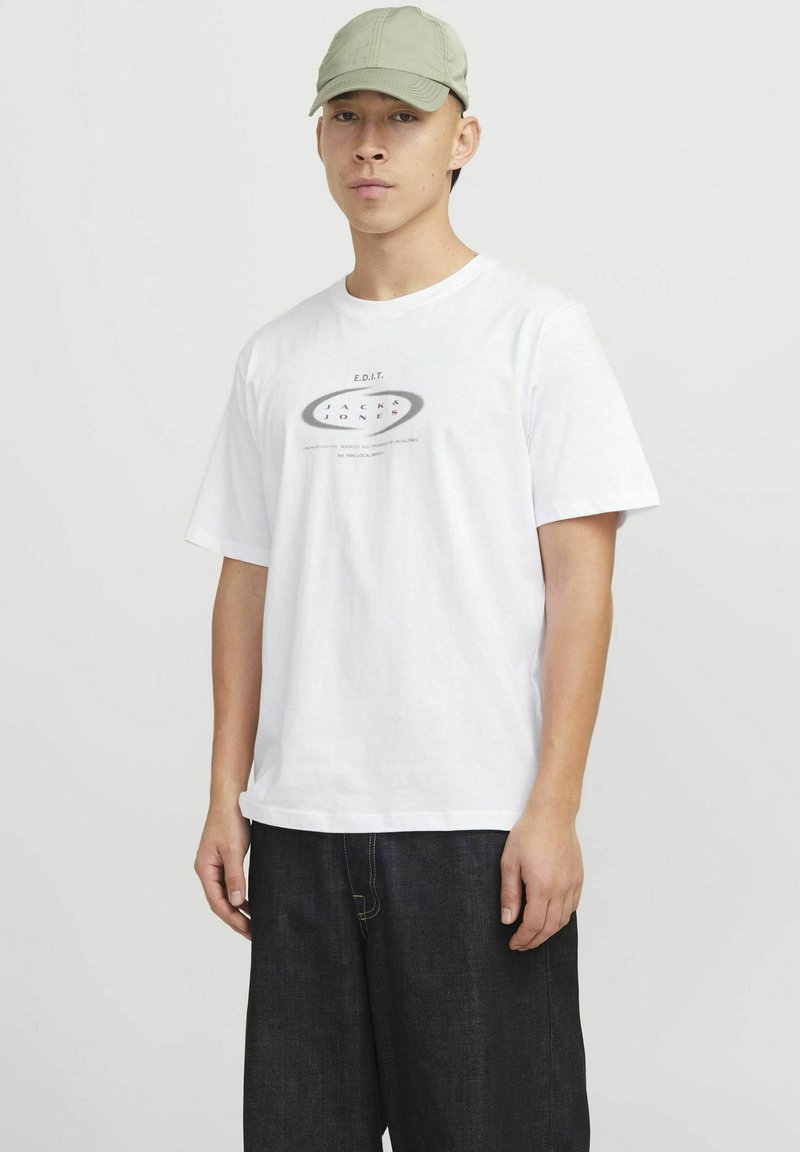 Hvid bomulds T-shirt med et trykt logo i grå og sort. Sammenstylet med mørke denimbukser. Modellen bærer en lysegrøn kasket.