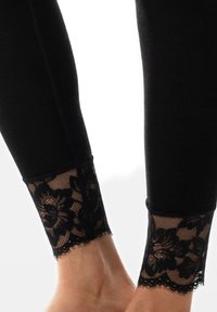 Leggings noirs à côtes avec une bordure en dentelle transparente au niveau de la cheville, ornés de motifs floraux et d'un bord festonné. Texture lisse sur l'ensemble.