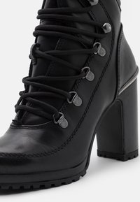 Bottine noire en cuir à talon haut avec talon bloc épais, bout rond, œillets métalliques et lacets noirs croisés.