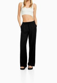 Svarta byxor med vida ben och dragsko i midjan, ihop med en vit ribbad croptop. Modellen står med händerna i fickorna och har på sig svarta sandaler.