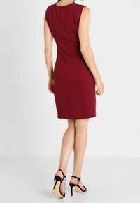 Vínové šaty bez rukávů s přiléhavým střihem, délkou ke kolenům a zadním zipem. Nošeny s černými sandály na vysokém podpatku se stahovacími pásky.