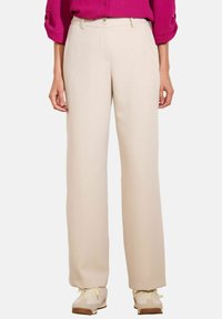 Vrouw in beige wijde broek, beige sneakers en een magenta langemouwen top met opgerolde mouwen, staande op een witte achtergrond.