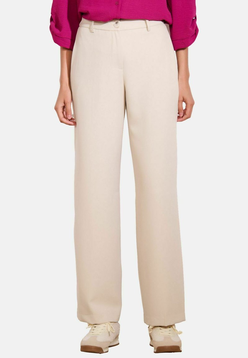 Vrouw in beige wijde broek, beige sneakers en een magenta langemouwen top met opgerolde mouwen, staande op een witte achtergrond.